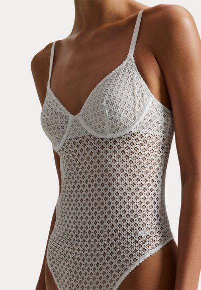 Γυναίκα που φοράει λευκό ολόσωμο κορμάκι από δαντέλα χωρίς μανίκια, με τριγωνικά κάπες και λεπτούς ρυθμιζόμενους ιμάντες, πάνω σε απλό φόντο.