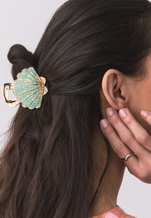Das dunkle Haar einer Frau ist teilweise mit einer gold- und türkisfarbenen muschelförmigen Haarspange zurückgesteckt, eine Hand mit einem goldenen Ring ruht an der Wange.