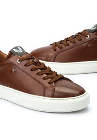 Sneakers in pelle marrone con suole in gomma bianca spessa e lacci marroni, caratterizzati da un piccolo logo metallico sul lato e fasce per il marchio sulla linguetta.