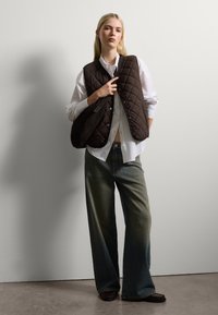 Gilet trapuntato marrone indossato sopra una camicia bianca, abbinato a jeans svasati sbiaditi. I materiali texturizzati e i colori contrastanti sono predominanti.
