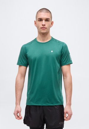 Giovane uomo con capelli corti che indossa una maglietta verde da atleta e pantaloncini neri, in piedi davanti a uno sfondo neutro.