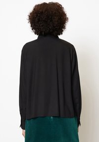 Schwarze Bluse mit langen Ärmeln, die einen Kragen hat, aus glattem Stoff und mit geknöpften Manschetten, kombiniert mit einem grünen Cordrock.