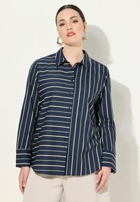 Camicia a maniche lunghe blu navy con strisce verticali crema, abbottonatura frontale, colletto a punta e una tasca sul petto. Tessuto dalla trama liscia.