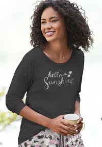 Crna majica dugih rukava s otisnutim natpisom "hello Sunshine", ležeran kroj, u kombinaciji s uzorkastim pidžama hlačama. Model drži šalicu.