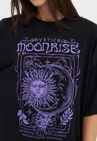 Black cotton t-shirt featuring a purple illustrated design with a sun, moon, and floral accents, text reads "Mystical Moonrise."
