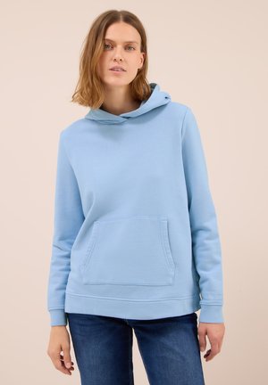 Femme portant un sweat à capuche bleu clair avec une poche frontale et un jean bleu foncé, se tenant devant un fond beige.