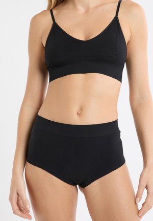 Schwarzes Bustier mit dünnen Trägern, das eine gerippte Textur und einen glatten Bund aufweist. Kombiniert mit hoch sitzenden schwarzen Unterteilen ähnlicher Textur.