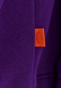 Fialová látka s hladkým povrchem obsahuje oranžovou etiketu s grafikou slunce. Barva je živá a přispívá k celkovému designu.