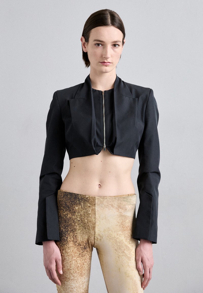 Black cropped jacket with long sleeves and front zipper, paired with fitted beige leggings featuring a textured gold pattern.