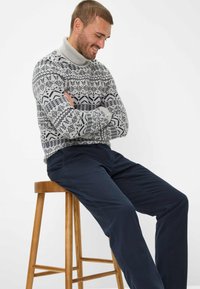 Maglione lavorato a maglia grigio a fantasia con colletto alto, abbinato a pantaloni blu navy. Sotto, uno sgabello di legno, individuo seduto con le braccia incrociate.
