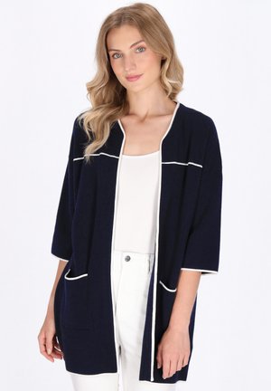 Marineblaue Strickjacke mit kurzen Ärmeln, weißem Besatz und zwei Fronttaschen. Weiche Textur und lockere Passform, ideal zum Überziehen über Outfits.