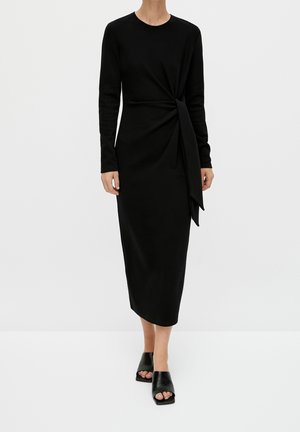 Kvinde iført en sort midi-kjole med lange ærmer og en sidesløjfe, kombineret med sorte åbne hæle sko, stående foran en ensfarvet baggrund.