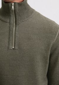 Khaki-grüner Reißverschluss-Pullover mit hohem Kragen. Strukturierter Strickstoff und ein metallischer Reißverschluss am Hals. Leichte Konstruktion.