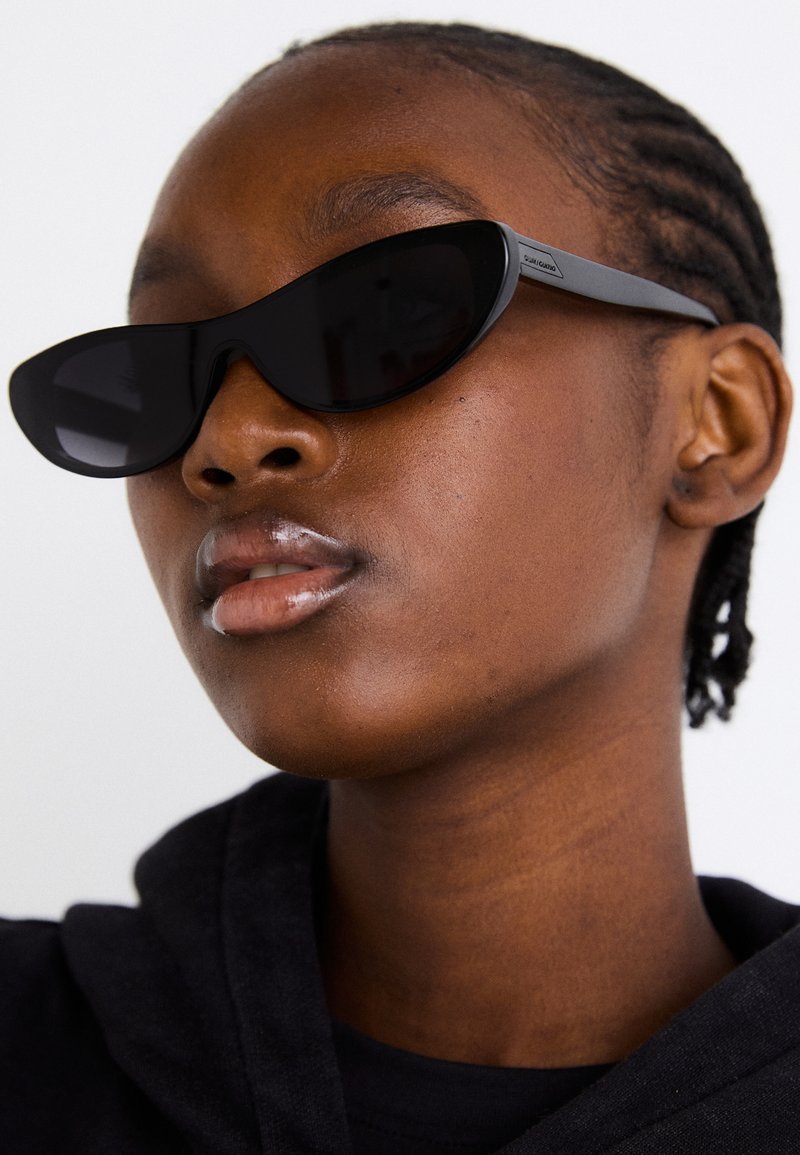Czarne okulary przeciwsłoneczne w stylu cat-eye z matowym wykończeniem, charakteryzujące się eleganckim designem i delikatnie zaokrąglonymi krawędziami, na tle ciemnego ubrania modelki.