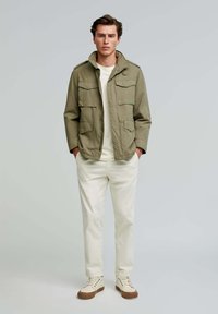 Olive green jacket with multiple front pockets, worn over a light-colored shirt and white pants. Beige sneakers complete the outfit.