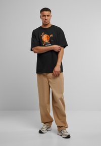 Svart bomullströja med orange basketgrafik, avslappnad passform, kombinerat med ljusa beige byxor och grå sneakers, står framför en enfärgad bakgrund.