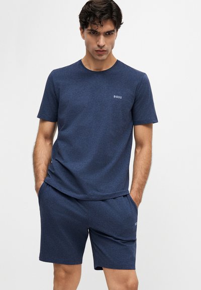 Homme portant un t-shirt à manches courtes bleu foncé et un short assorti avec le logo "BOSS", debout, mains dans les poches, devant un fond uni.