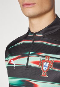 Zwart sportshirt met groene en rode abstracte strepen, ritskraag, Puma-logo en het embleem van het Portugese nationale team op de borst.
