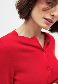 Punane kootud cardigan, millel on laineline kaelus, kaks nuppu ja soonikkoes. Suum vaade rõhutab materjali ja disainielemente.