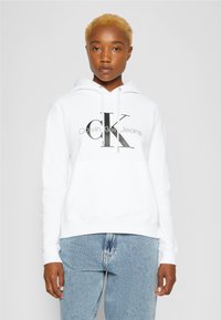 Felpa bianca realizzata in tessuto morbido, con un logo nero "CK" e "Calvin Klein Jeans" in grigio. Vestibilità casual e rilassata con cappuccio regolabile.