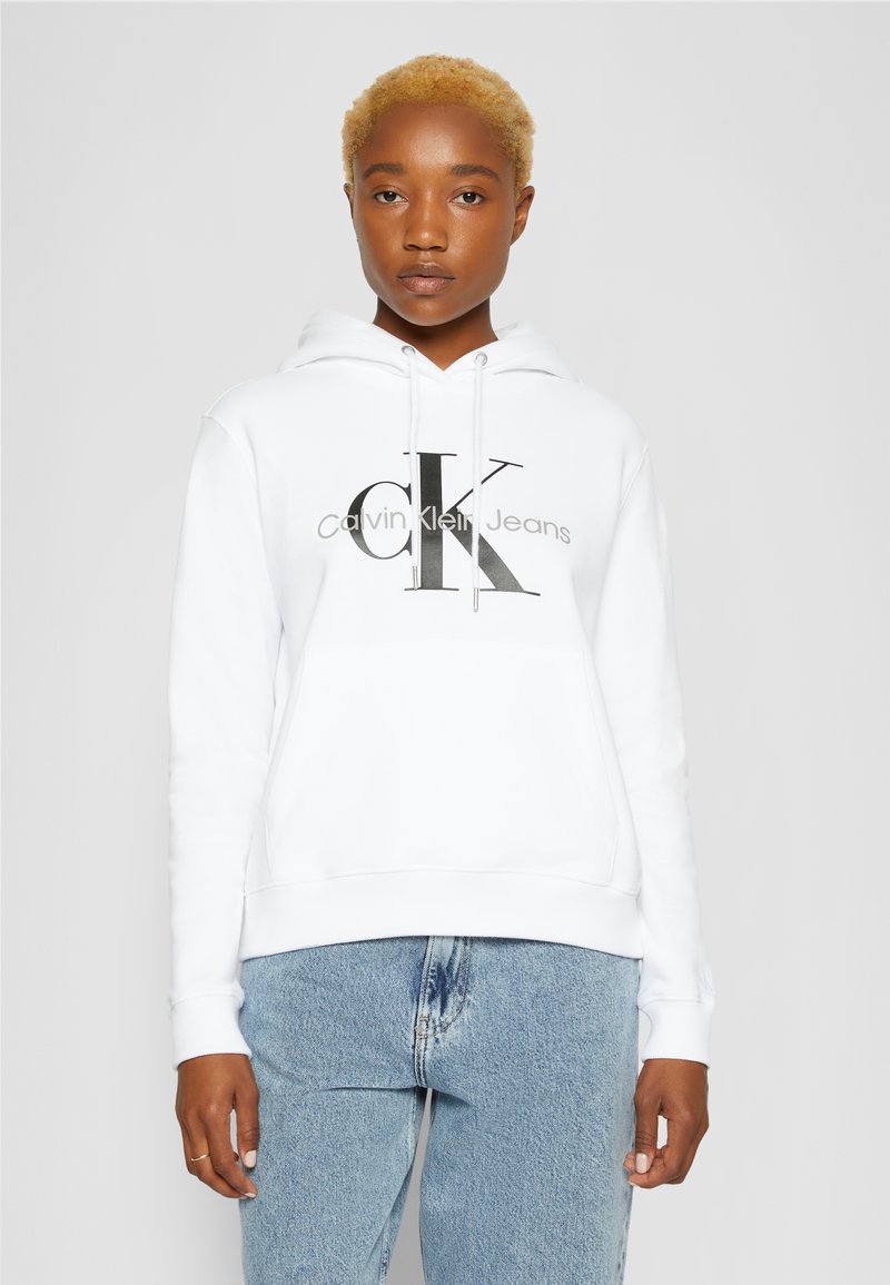 Felpa bianca realizzata in tessuto morbido, con un logo nero "CK" e "Calvin Klein Jeans" in grigio. Vestibilità casual e rilassata con cappuccio regolabile.