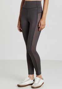 Femme portant des leggings taille haute gris foncé avec des panneaux en filet sur les côtés et des baskets blanches, se tenant sur une surface gris clair.
