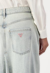 Jeans de mezclilla azul claro con un parche triangular rojo y blanco del logo "GUESS?" en el bolsillo trasero. La tela presenta una textura ligeramente desgastada.