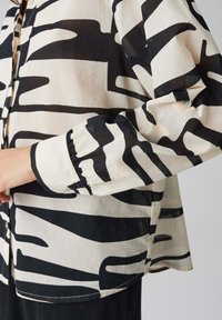 Blusa nera e crema a fantasia con vestibilità ampia, maniche lunghe, dettagli di bottoni e polsini arricciati che mostrano linee astratte e ondulate.
