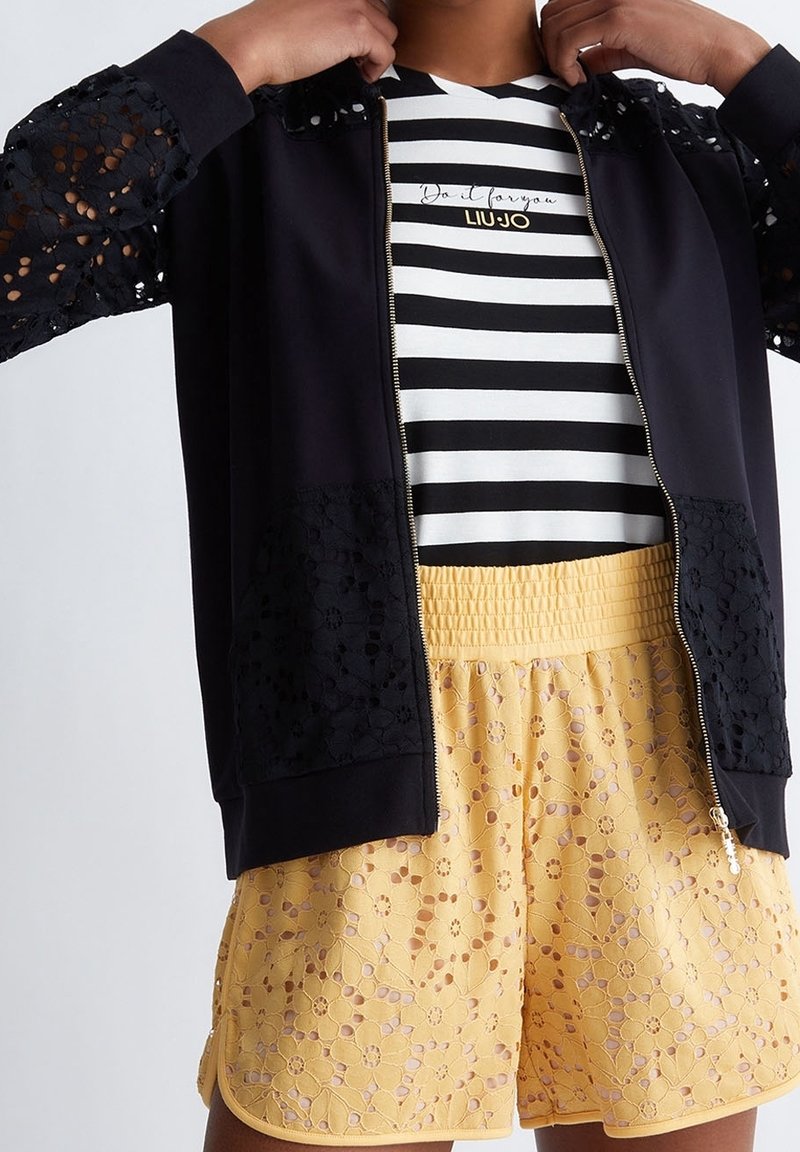 Černá zipová bunda s krajkovými detaily na rukávech a kapsách, spárovaná se žlutými krajkovými šortkami s květinovými vzory a pruhovaným tričkem.