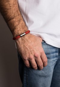 Raudonas pintas apyrankė su sidabro spalvos metaline sagtimi, dėvima ant riešo. Rankos pirštai remiasi į džinsus, rodančius denimų tekstūrą ir toną.