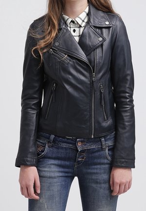 Femme portant une veste en cuir noire de moto par-dessus une chemise à carreaux noire et blanche et un jean bleu, debout les bras détendus le long du corps.