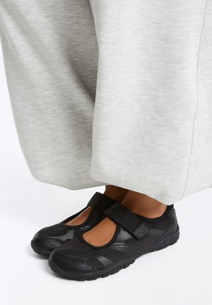 Pieds portant des chaussures Mary Jane noires en mesh et cuir avec des bandes Velcro, associées à un pantalon large gris clair.