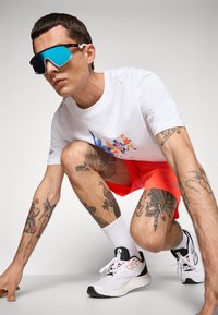 Mann som croucher, iført blå speilbriller, hvit t-skjorte med grafikk, lys røde shorts, hvite sokker, hvite sneakers og synlige tatoveringer på armer og ben.