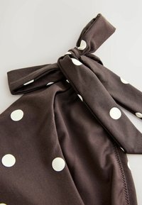 Brūna auduma ar baltām punktiņām, ar sasietu loku dekorāciju. Gluda tekstūra, ar slāņotu dizainu un redzamām šuvēm gar malām.