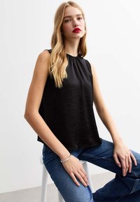 Top senza maniche nero con scollatura arricciata, realizzato in tessuto liscio. Il modello lo abbina a jeans di denim blu seduto su uno sgabello bianco.
