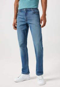 Jeans in denim blu con taglio dritto, cinque tasche e leggera scoloritura. Abbinati a sneakers bianche, su uno sfondo neutro.