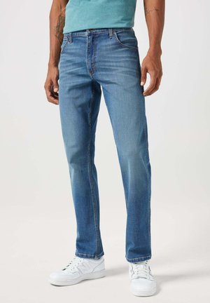 Jeans in denim blu con taglio dritto, cinque tasche e leggera scoloritura. Abbinati a sneakers bianche, su uno sfondo neutro.