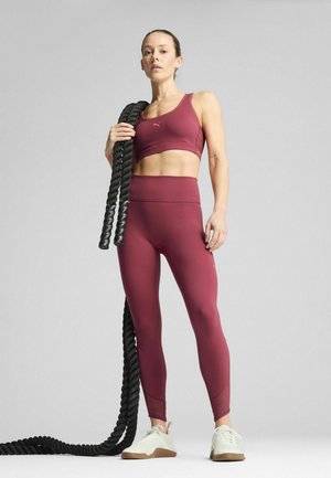 Sportovní souprava se bordo crop topem a legínami s vysokým pasem, zkombinovaná s bílými a béžovými teniskami, držící černé cvičební lana.