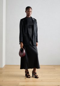 Svart skreddersydd blazer over en tettsittende svart kjole, kombinert med åpne svarte hæler og en teksturert bordeaux skulderveske. Glatt stoff, elegant silhuett.
