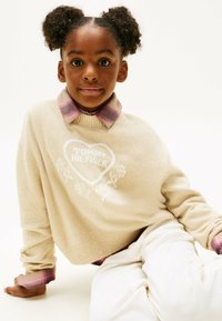 Beige Pullover mit weißem Herzlogo und floralem Design, übereinander getragen mit einem pinken Karohemd. Das Model trägt weiße Hosen. Weiche, strukturierte Strickware.