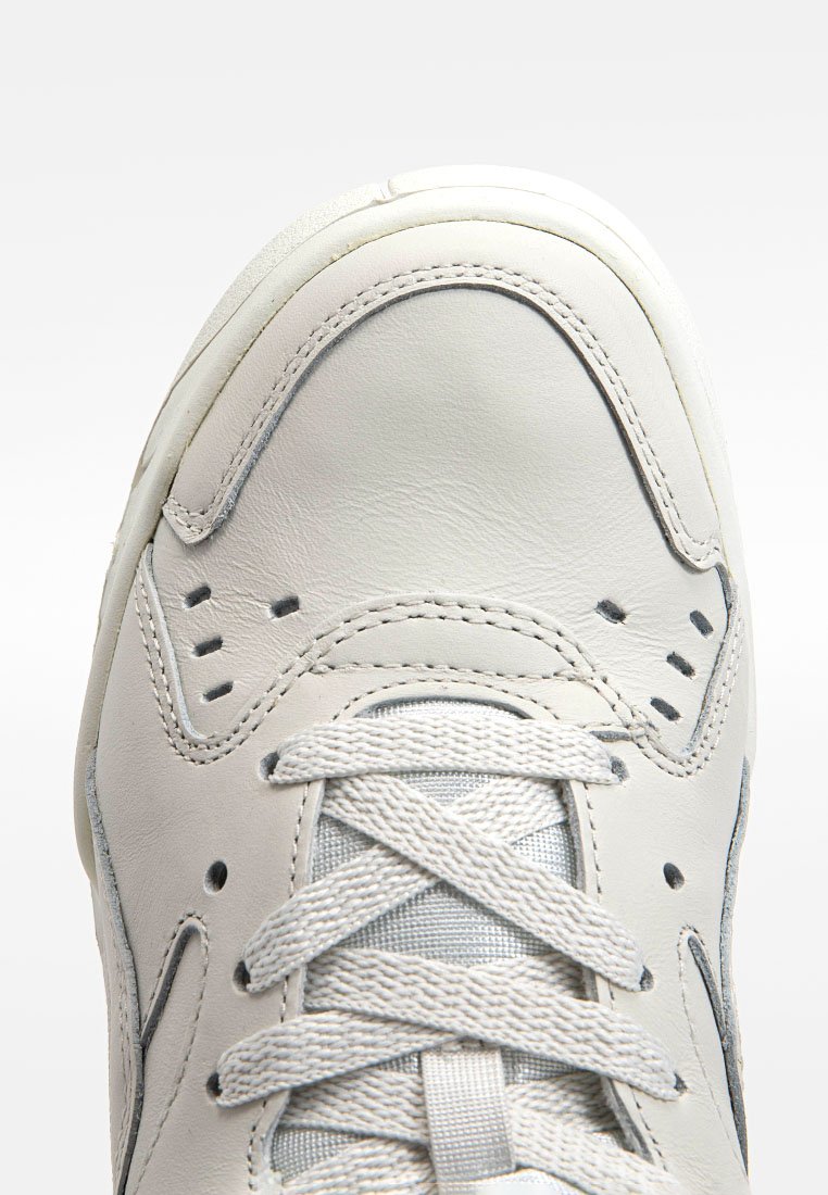 Sneaker en cuir blanc avec une tige lisse, des accents texturés, un col rembourré et des lacets gris. Le design comprend des perforations pour la respirabilité.
