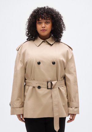 Trench-coat beige à double boutonnage, avec de gros boutons noirs et une taille ceinturée. Il présente un col pointu et des manches larges. Matière lisse.