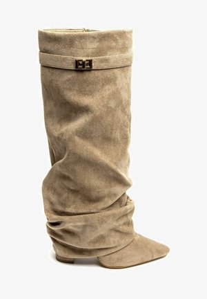 Botte beige en daim souple et ample, avec un bout pointu et une lanière à boucle rectangulaire dorée près du haut.