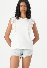 Top sin mangas de algodón blanco con hombros con encaje. Combinado con pantalones cortos de mezclilla desgastados, con dobladillos deshilachados y un ajuste casual.