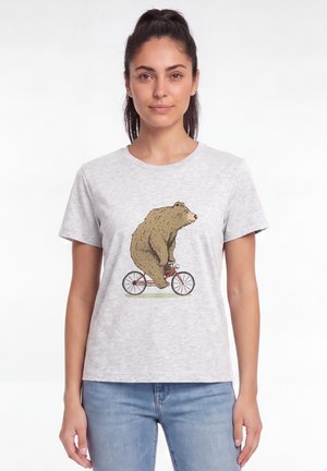 Femme portant un t-shirt gris clair avec un dessin d’un ours brun sur un vélo rouge, associé à un jean bleu.