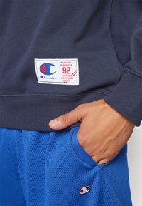 Sudadera navy con una etiqueta blanca que presenta acentos en rojo y azul, combinada con pantalones cortos deportivos de malla azules, destacando la transpirabilidad y la comodidad.