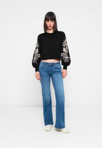 Suéter negro corto con bordado floral blanco en las mangas abullonadas, combinado con jeans de campana azul claro y zapatillas blancas. Fondo sencillo.