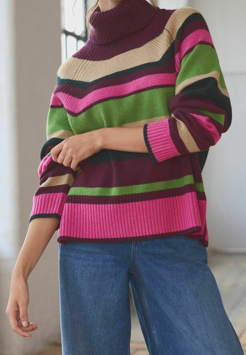 Personne portant un pull à col roulé côtelé à rayures roses, vertes, beiges et bordeaux, associé à un jean bleu, à l'intérieur près d'une fenêtre.