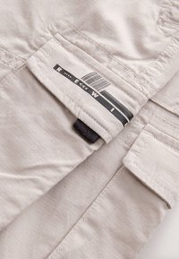 Tessuto beige chiaro con una sottile texture a coste verticali. Presenta un'etichetta con design a codice a barre in bianco e nero e un piccolo anello nero di attacco.