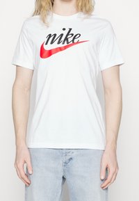 Valkoinen puuvillapaita, jossa on musta "nike"-logo ja punainen swoosh-muotoilu. Lyhyet hihat ja klassinen pyöreä kaula-aukko.